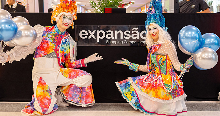 Dois personagens em frente a placa do evento de inauguração da expansão do Shopping Campo Limpo